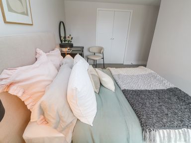 A bedroom with a bed with pillows and blankets a chair a bedside table with a lamp and flowers at Porth Beach in Porth near St Columb Minor