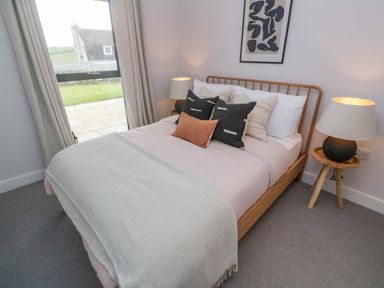 A bedroom with a wooden bed frame, pillows, a throw blanket, a bedside table with a lamp, a window with curtains and a framed artwork on the wall at Porth Beach in Porth near St Columb Minor