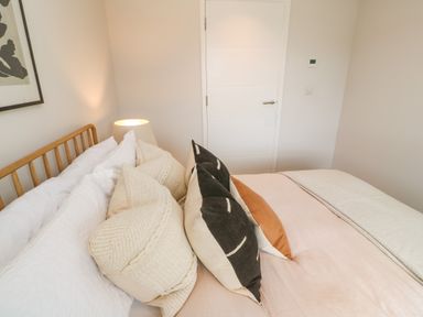 A bedroom with a wooden bed frame white and beige pillows a bedside lamp and a closed white door at Porth Beach in Porth near St Columb Minor