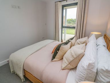 A bedroom with a bed covered with pillows and a blanket next to a window with curtains at Porth Beach in Porth near St Columb Minor