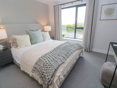 A bedroom with a bed with pillows and a blanket bedside tables with lamps a window with curtains and a framed picture on the wall at Porth Beach in Porth near St Columb Minor