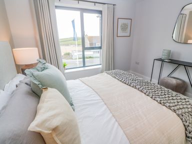A bedroom with a bed with pillows and a blanket a lamp on a bedside table a window with curtains a framed picture a mirror on the wall a console table with a decorative item and a stool at Porth Beach in Porth near St Columb Minor