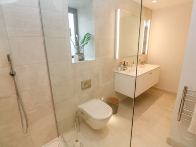 A bathroom with a wall-mounted toilet a glass shower enclosure a double sink vanity and a small window with a plant at Porth Beach in Porth near St Columb Minor