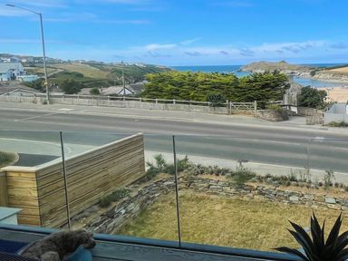 A coastal view with a road, green hills, beach, and ocean seen from behind a glass balcony with a dog lying down and a plant nearby at Porth Beach in Porth near St Columb Minor