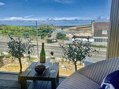 A balcony with a wooden table holding a bottle of champagne and two glasses overlooking a beach and road at Porth Beach in Porth near St Columb Minor