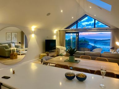 An open living and dining area with a dining table, chairs, a sofa, armchairs, a television, a large round wall mirror, and a window showing a coastal view at Porth Beach in Porth near St Columb Minor
