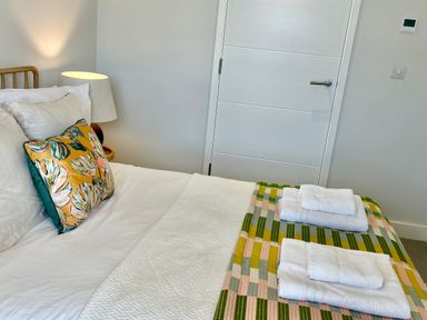 A bed with white linens and a colorful pillow next to a nightstand with a lamp at Porth Beach in Porth near St Columb Minor