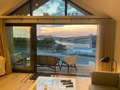 A living room with large glass sliding doors opening to a balcony with two chairs and a view of a beach and hills at Porth Beach in Porth near St Columb Minor