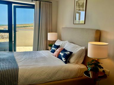 A bedroom with a bed with pillows and cushions next to a glass door with curtains and a lamp on a side table at Porth Beach in Porth near St Columb Minor