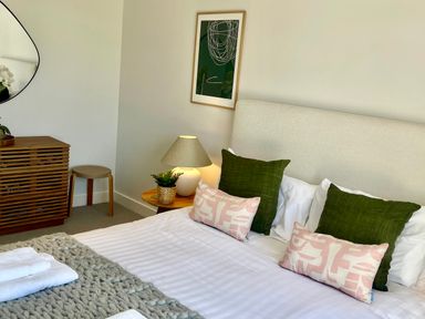 A bedroom with a bed with green and pink pillows a wooden nightstand with a lamp and plant a wooden dresser and a round mirror at Porth Beach in Porth near St Columb Minor