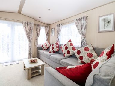 A living room with a grey sofa with red and white cushions a wooden coffee table and floral curtains at Freddy's Lodge Foxburrow Hang in Fritton