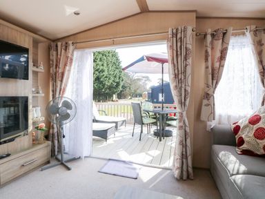 A living room with a sofa TV fan and sliding door opening to a patio with table and chairs at Freddys Lodge Foxburrow Hang in Fritton