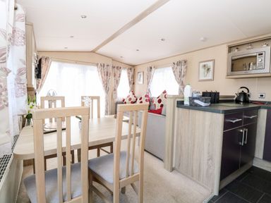 A dining area with a wooden table and chairs next to a living room with a sofa and cushions and a kitchen counter with a kettle and microwave at Freddy's Lodge Foxburrow Hang in Fritton