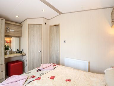 A bedroom with a bed covered with a floral bedspread towels on the bed a dressing table with a mirror and hairdryer two closed wooden doors and a red ottoman at Freddy's Lodge Foxburrow Hang in Fritton