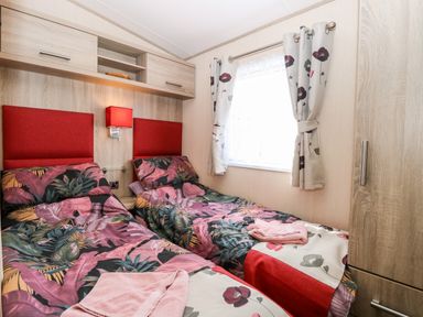 A bedroom with two single beds with floral bedding red headboards a window with floral curtains and wooden cabinets at Freddys Lodge Foxburrow Hang in Fritton