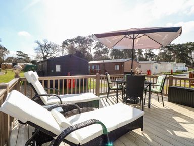 A deck with two lounge chairs and a table with an umbrella overlooking cabins and trees at Freddy's Lodge Foxburrow Hang in Fritton