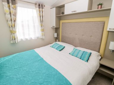 A bedroom with a bed blue bedspread teal pillows window with patterned curtains and shelves at Moonlight Lodge Bockenfield Country Holiday Park near West Thirston