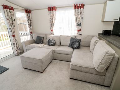 A living room with a gray sectional sofa, round and square cushions, floral curtains, and a sliding glass door at Moonlight Lodge in Bockenfield Country Holiday Park near West Thirston