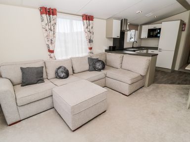 A living room with a beige sectional sofa and ottoman adjacent to a kitchen with white cabinets at Moonlight Lodge in Bockenfield Country Holiday Park near West Thirston
