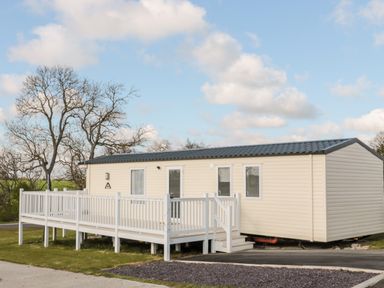 A beige mobile home with a white deck and railing on grass with leafless trees in the background at Moonlight Lodge in Bockenfield Country Holiday Park near West Thirston