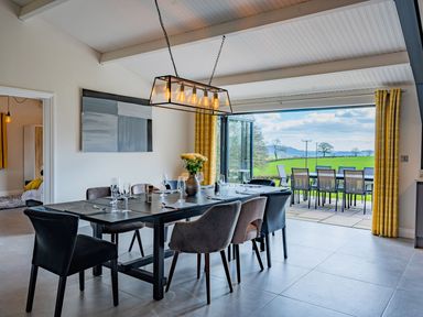 A dining room with a black table and mixed chairs next to open doors leading to a patio with outdoor chairs and tables at Quantock Barn in Stockland near Honiton
