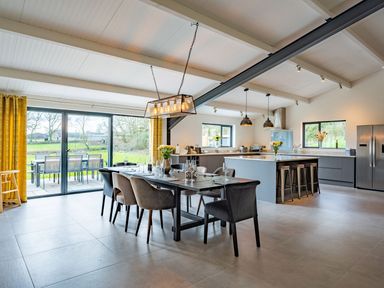 An open dining and kitchen area with a dining table set with glasses and chairs in front of large glass doors and a kitchen island with stools at Quantock Barn Stockland near Honiton