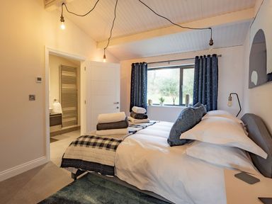 A bedroom with a bed, pillows, folded towels, a window with curtains, hanging light bulbs, and an open door leading to a bathroom at Quantock Barn in Stockland near Honiton