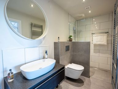 A bathroom with a sink, a round mirror, a wall-mounted toilet, a towel rack, and a walk-in shower at Quantock Barn in Stockland near Honiton