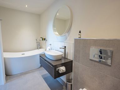 A bathroom with a freestanding bathtub a wall-mounted sink with a round mirror and a tiled wall at Quantock Barn in Stockland near Honiton