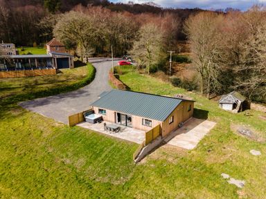 A single-story wooden house with a green roof and outdoor patio in a grassy area near a forest at Quantock Barn Stockland near Honiton