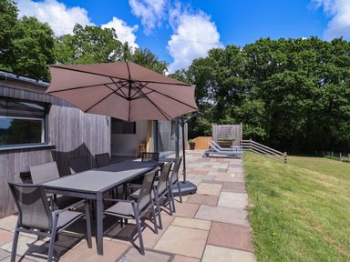 An outdoor patio with a dining table, chairs, a large umbrella, and two sun loungers next to a grassy area by Quantock Barn in Stockland near Honiton