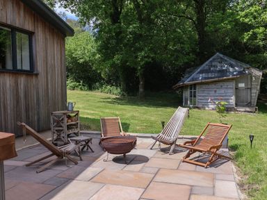 A patio with wooden lounge chairs and a fire pit next to a wooden building and a small shed at Quantock Barn in Stockland near Honiton