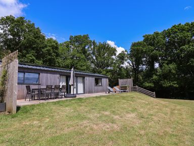 A wooden cabin with outdoor dining table and chairs and sun loungers on the deck surrounded by grass and trees at Quantock Barn in Stockland near Honiton