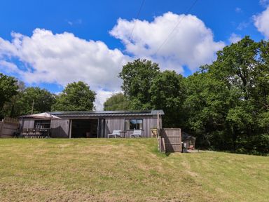 A detached wooden house with a patio and outdoor chairs surrounded by trees at Quantock Barn in Stockland near Honiton