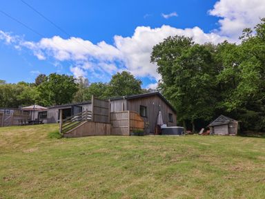 A wooden house with patio furniture and a small wooden shed on a grassy hill surrounded by trees at Quantock Barn in Stockland near Honiton