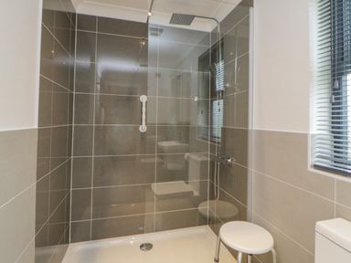 A shower area with a glass door a stool tiled walls and a window with blinds in a bathroom at Meadow Lodge in Lifton