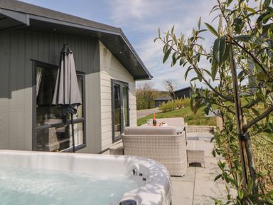 An outdoor patio area with a hot tub a table with chairs and a bottle of wine next to a modern house at Meadow Lodge in Lifton