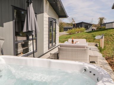 An outdoor patio with a hot tub wicker seating table umbrella and houses on a grassy hill at Meadow Lodge in Lifton