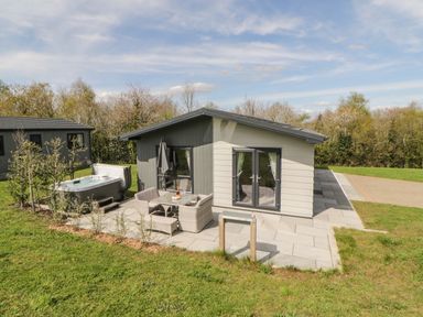 A modern single-story cabin with outdoor seating and a hot tub on a patio surrounded by grass and trees at Meadow Lodge in Lifton