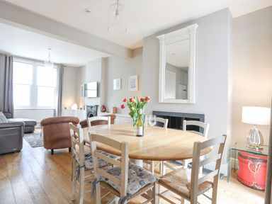 A dining area with a wooden table and six chairs with cushions next to a living area with seating a TV and a window at Cobblers Cottage in Beaumaris