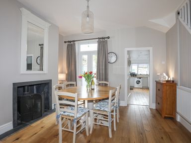 A dining room with a round wooden table surrounded by six chairs with patterned cushions a fireplace a mirror on the wall and a wooden cabinet near a doorway to a kitchen at Cobblers Cottage in Beaumaris