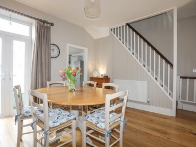 A dining area with a round wooden table and six chairs with cushions a vase with flowers on the table a staircase with white railings and wooden banister a clock on the wall and a wooden floor at Cobblers Cottage in Beaumaris