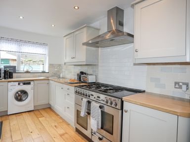 A kitchen with white cabinets wooden countertops a washing machine a gas stove and oven and a window with a blind at Cobblers Cottage in Beaumaris