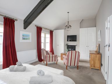 A bedroom with a bed and rolled towels red striped armchairs a wall mounted tv and red curtains at Cobblers Cottage in Beaumaris