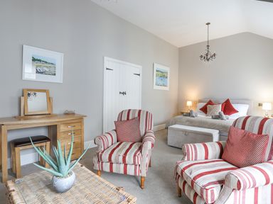 A bedroom with a bed, two lamps, striped armchairs, a desk with a mirror, and a plant on a wicker table at Cobblers Cottage in Beaumaris