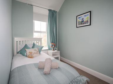 A small bedroom with a single bed with pillows and a teddy bear a white nightstand with a colorful lamp and a framed picture on the wall at Cobblers Cottage in Beaumaris