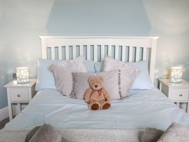 A bedroom with a white bed frame blue bedding a teddy bear and two bedside tables with lamps at Cobblers Cottage in Beaumaris