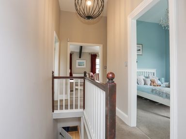 A hallway with a wooden banister and doors leading to a bedroom with a white bed frame and a living room with red curtains at Cobblers Cottage Beaumaris