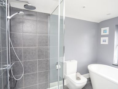 A bathroom with a glass shower enclosure a white toilet with a towel on top and a freestanding bathtub with two pictures on the wall at Cobblers Cottage in Beaumaris