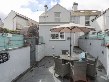 A patio area with a round glass table and six wicker chairs under a large umbrella at Cobblers Cottage in Beaumaris
